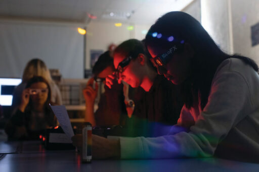 Students use diffraction grating glasses to observe the separated frequencies of light from gas discharge lamps in order to determine the elemental composition of the gas.